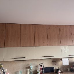 MatWood Design - Dwukolorowe szafki kuchenne: górne z drewnopodobnym frontem, dolne kremowe z podłużnymi uchwytami. Widoczny fragment blatu i wyposażenia kuchennego.