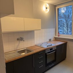 MatWood Design - Nowoczesna, kompaktowa kuchnia z czarnymi szafkami i drewnianym blatem. Widoczny zlew, płyta grzewcza i piekarnik. Białe płytki na ścianie i okno z widokiem na drzewa.