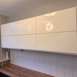 MatWood Design - Białe, lakierowane szafki kuchenne wiszące nad blatem z drewnopodobnym dekorem i białymi płytkami. Widoczne odbicie światła na frontach.