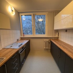 MatWood Design - Kuchnia z czarnymi szafkami i drewnianym blatem, widok na okno z drzewami. Białe płytki na ścianie i okrągła lampa na suficie. Ujęcie z perspektywy podłogi.