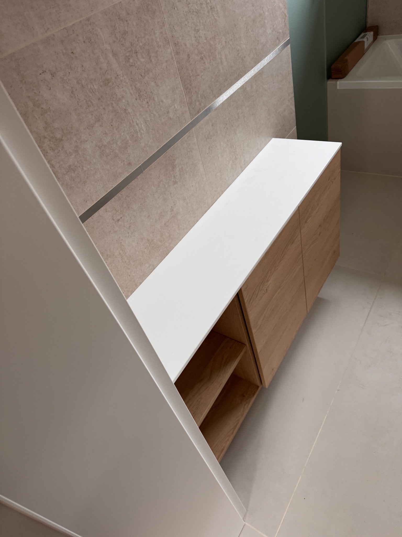 Nowoczesna szafka łazienkowa z drewnianymi frontami i białym blatem, wkomponowana w beżową ścianę z metalowym akcentem. Widok z góry, minimalistyczny design.