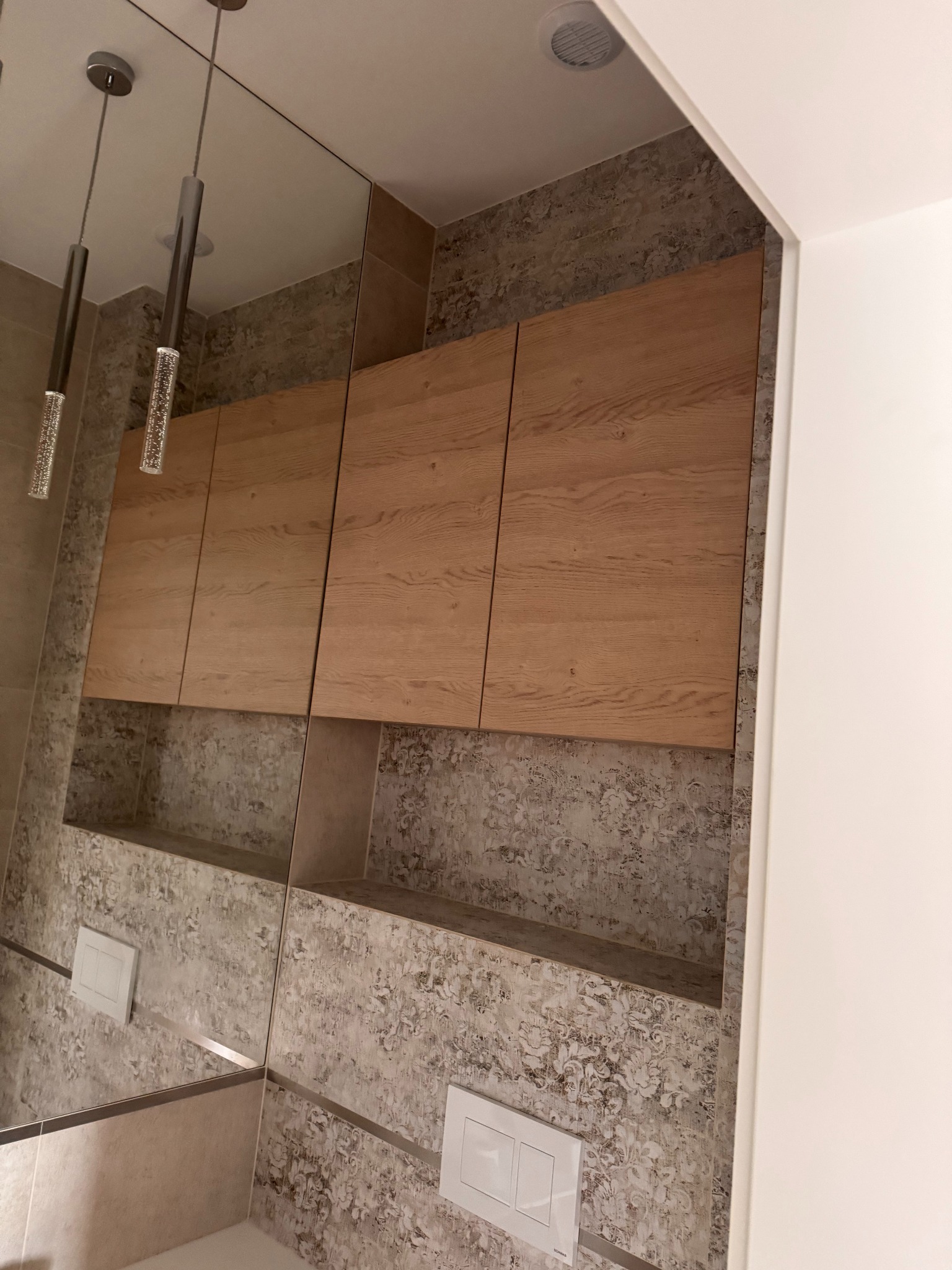 Drewniane szafki wiszące nad zabudowaną półką w łazience z lustrem i dekoracyjną tapetą. Nowoczesne oświetlenie punktowe.