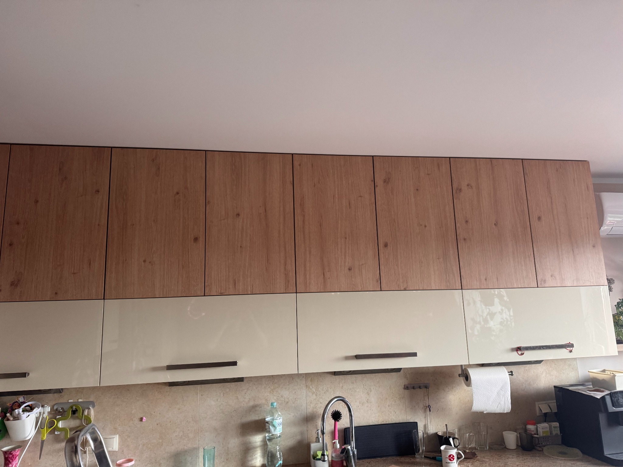 Dwukolorowe szafki kuchenne: górne z drewnopodobnym frontem, dolne kremowe z podłużnymi uchwytami. Widoczny fragment blatu i wyposażenia kuchennego.