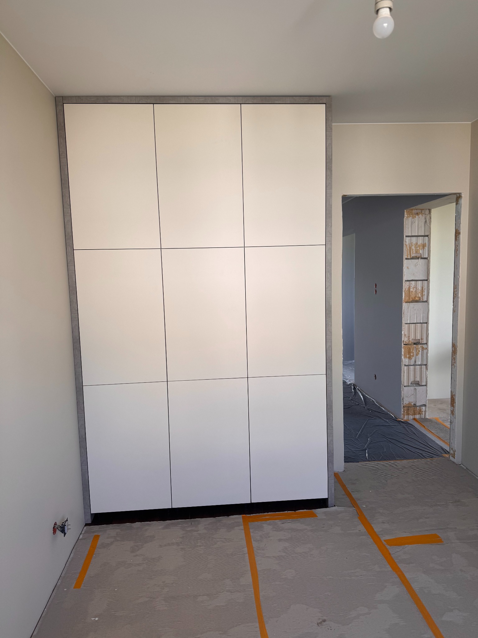 Biała szafa wnękowa z szarym obramowaniem, widoczna w surowym wnętrzu z przygotowanym podłożem pod podłogę i otwartym przejściem do innego pomieszczenia.