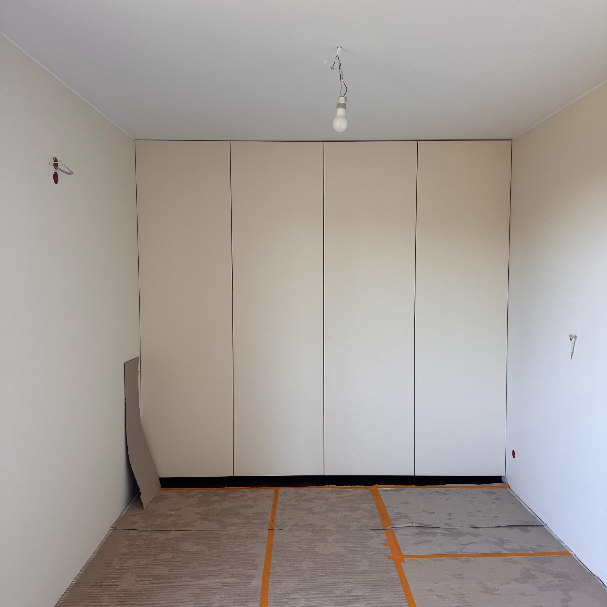 Jasna szafa wnękowa z czterema drzwiami w pokoju w trakcie wykańczania. Podłoga zabezpieczona, widoczna żarówka na kablu. Minimalistyczny design.