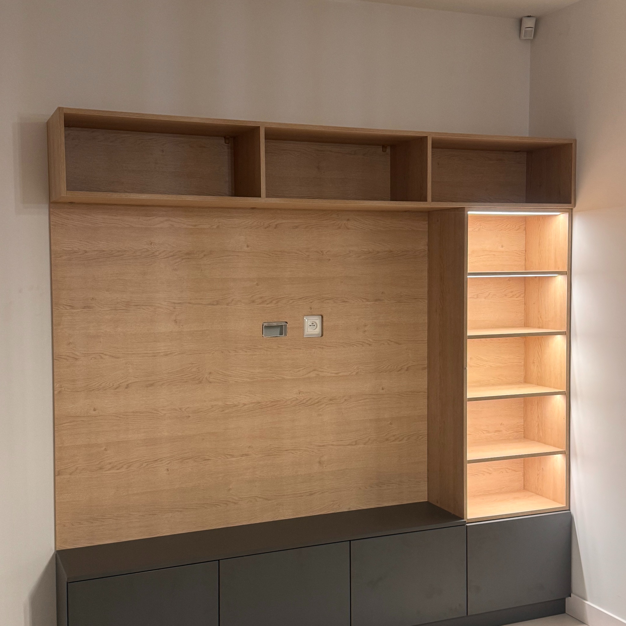 Drewniana zabudowa ściany z szafkami górnymi i dolnymi w kolorze grafitowym. Półki z podświetleniem LED. Gniazdka elektryczne na drewnianym panelu.