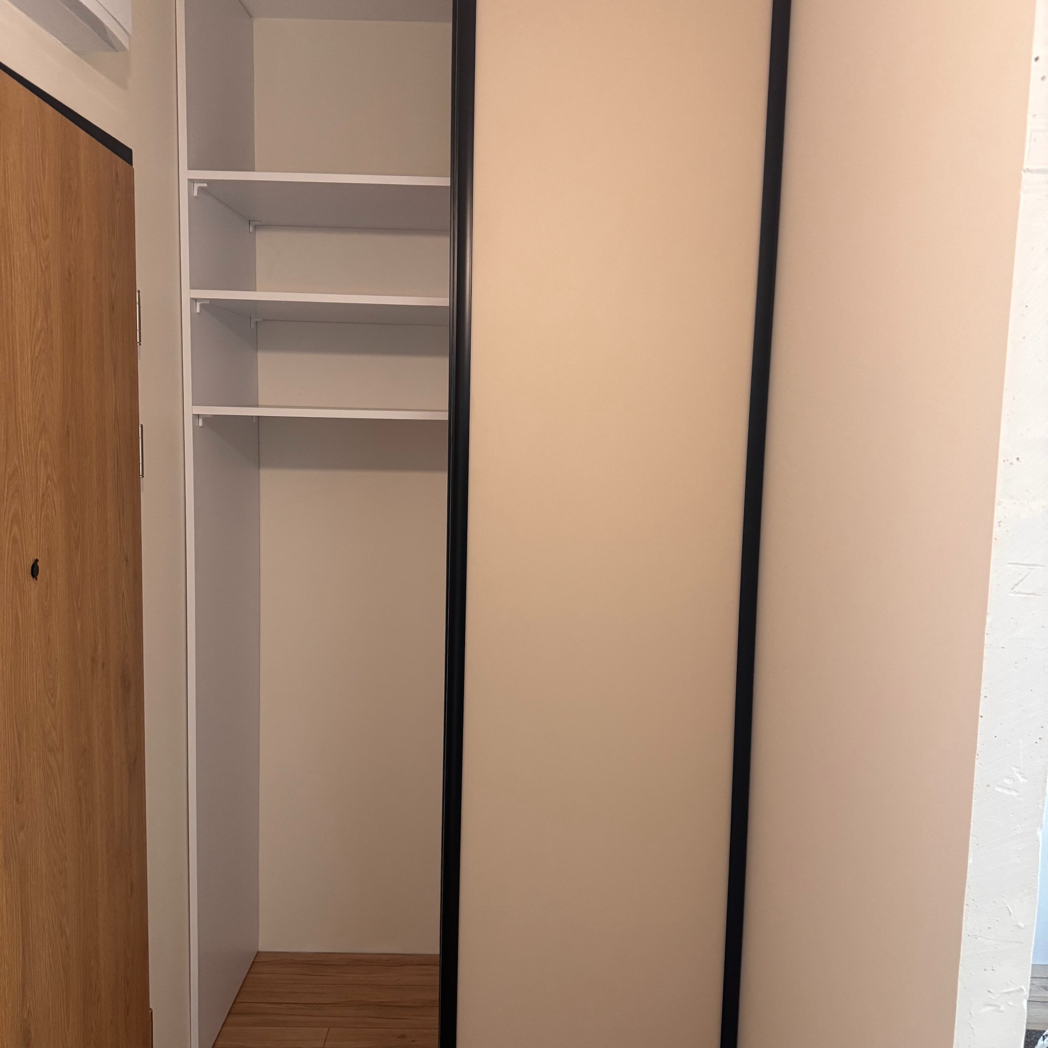 Jasna szafa wnękowa z przesuwnymi drzwiami w odcieniach beżu i czerni, z widocznymi półkami wewnątrz. Minimalistyczny design, drewniana podłoga.