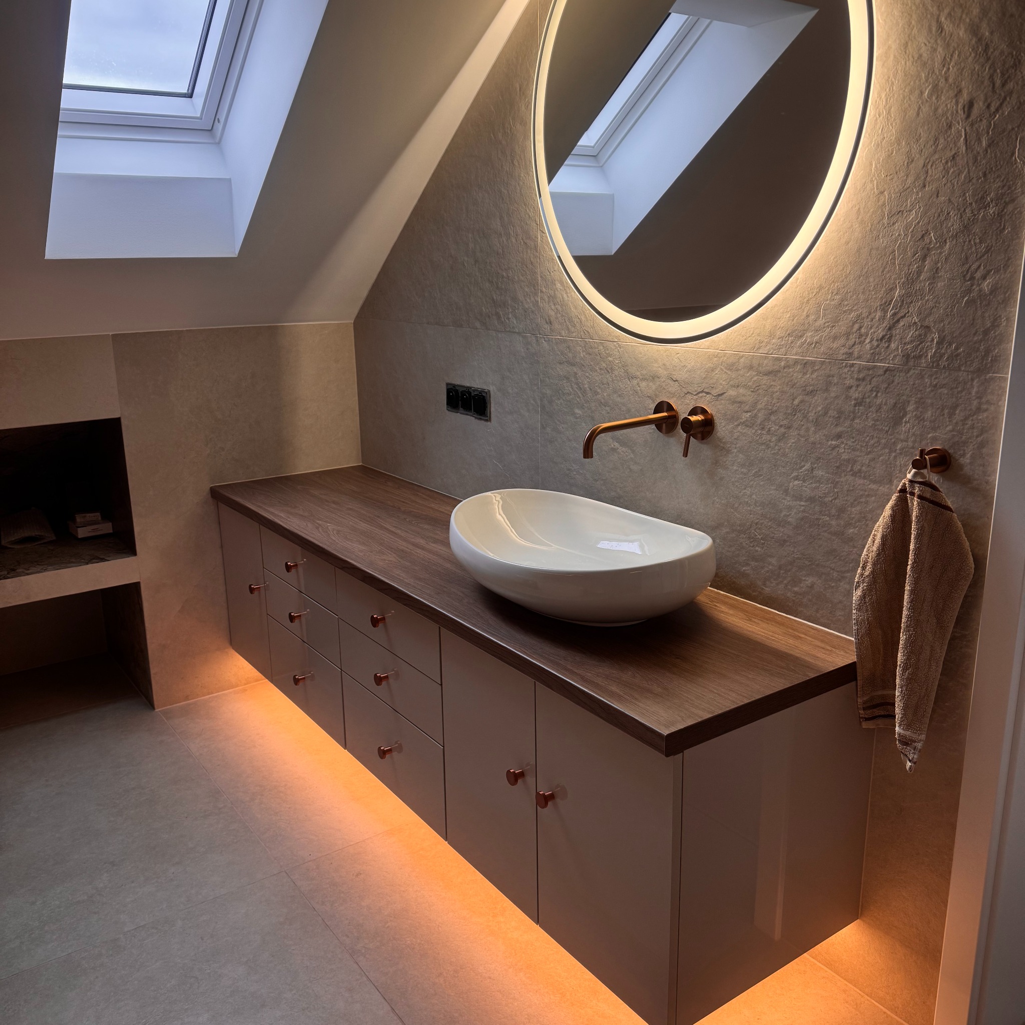 Łazienka na poddaszu z szafką pod umywalkę, blatem z ciemnego drewna i podświetleniem LED. Okrągłe lustro z oświetleniem i skośny sufit z oknem dachowym.