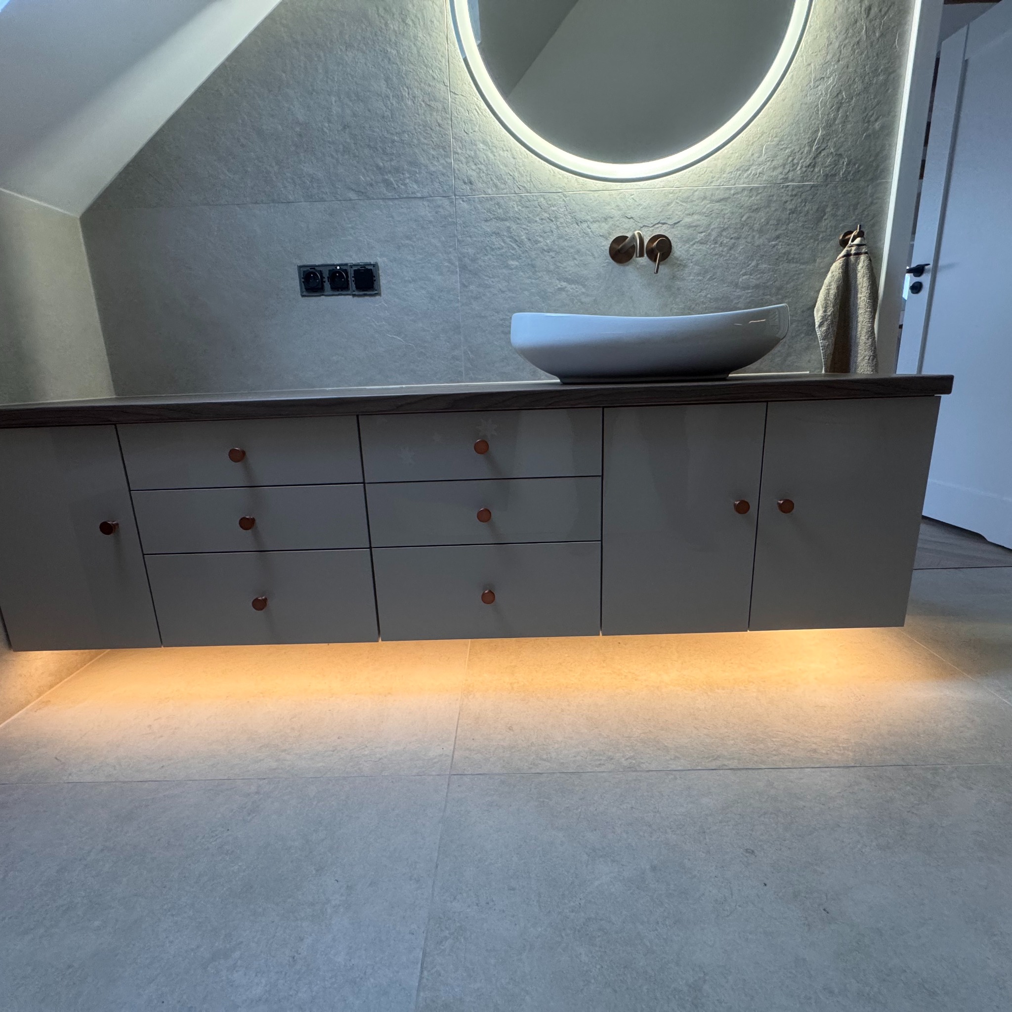 Nowoczesna szafka łazienkowa z szufladami i oświetleniem LED pod spodem, umywalka nablatowa i okrągłe lustro z podświetleniem. Minimalistyczny design.