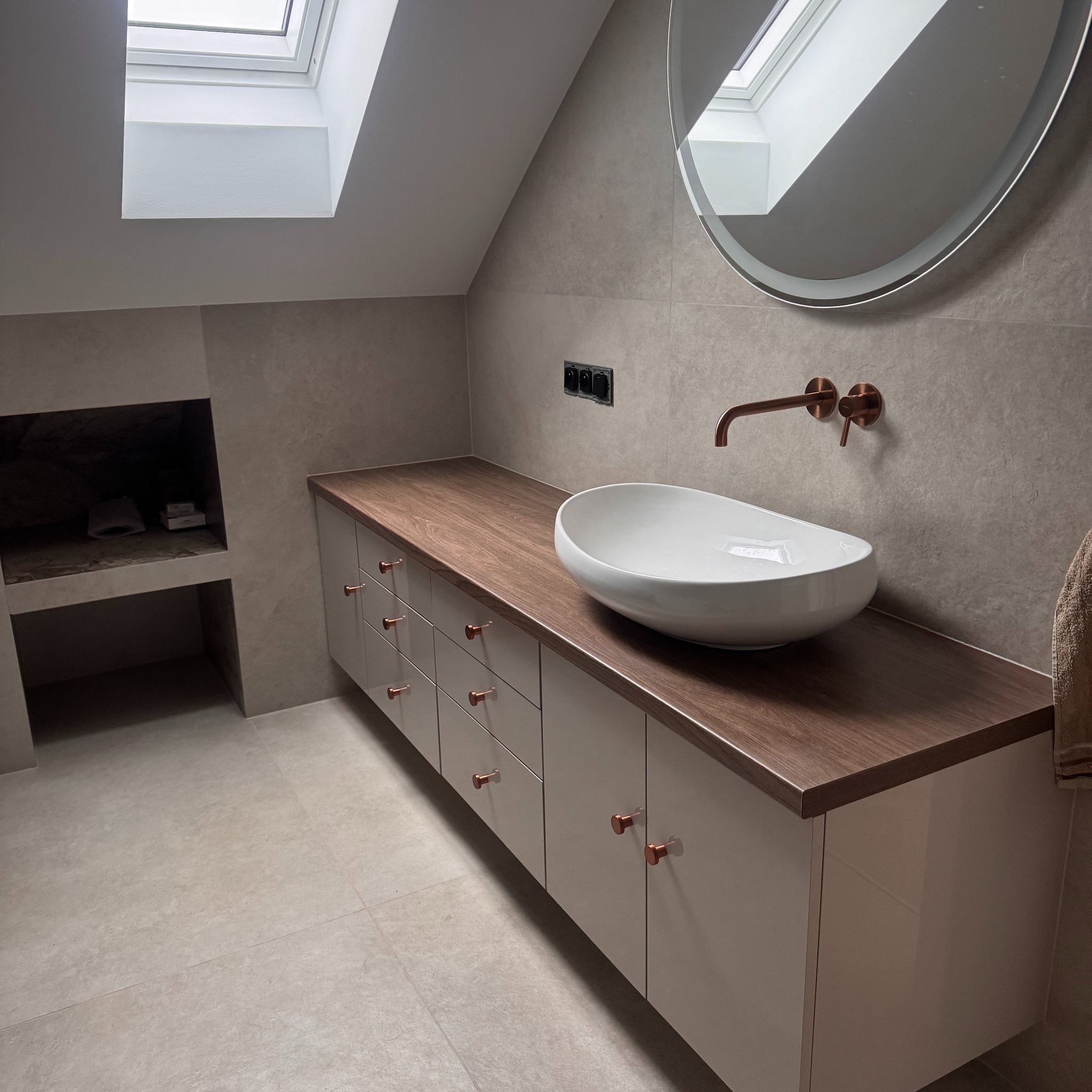 Nowoczesna łazienka na poddaszu z szafką pod umywalkę z drewnianym blatem i miedzianą armaturą. Okrągłe lustro z podświetleniem i okno dachowe.