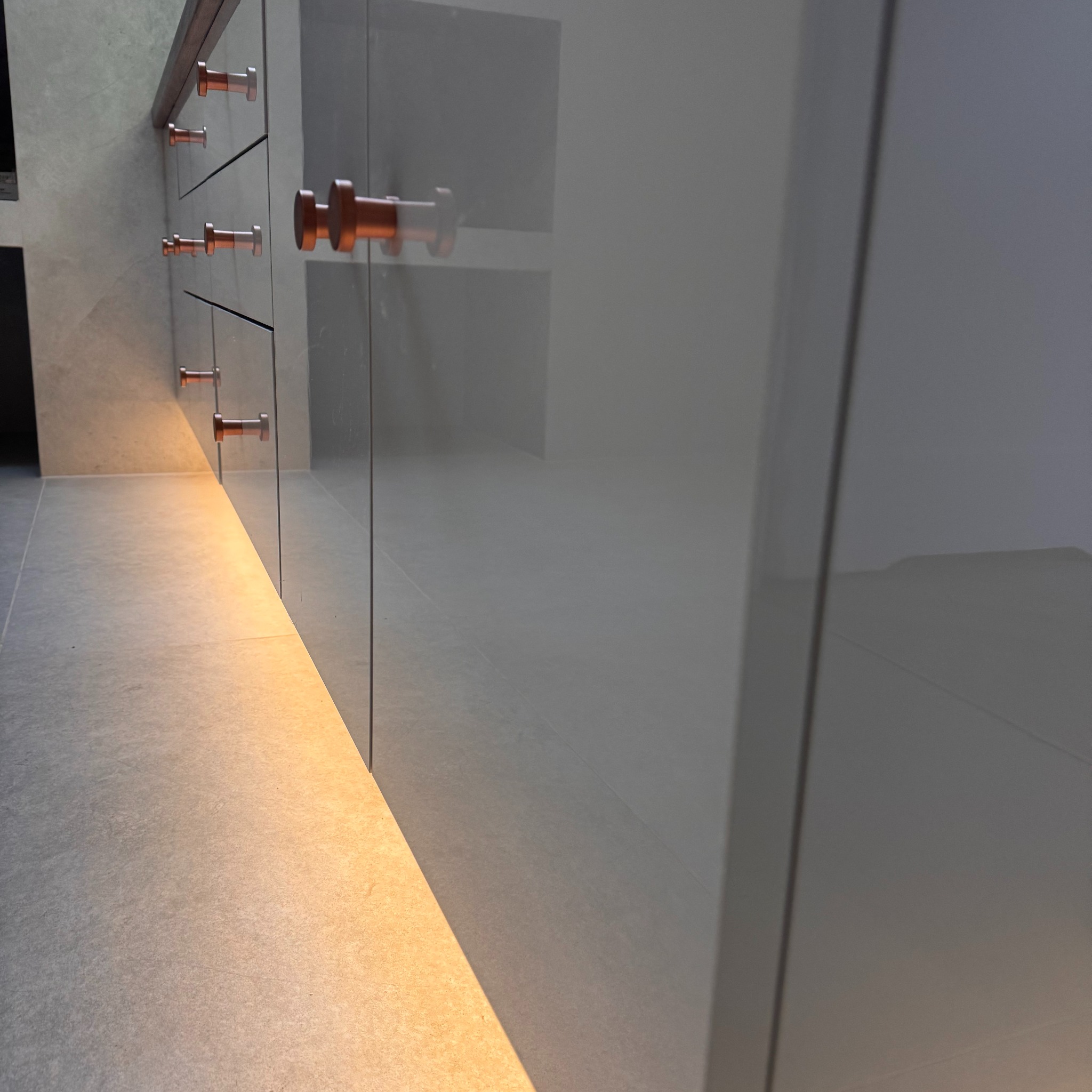 Szare, lakierowane fronty szafek z miedzianymi uchwytami, oświetlone od dołu ciepłym światłem. Minimalistyczny design, nowoczesne wykończenie.