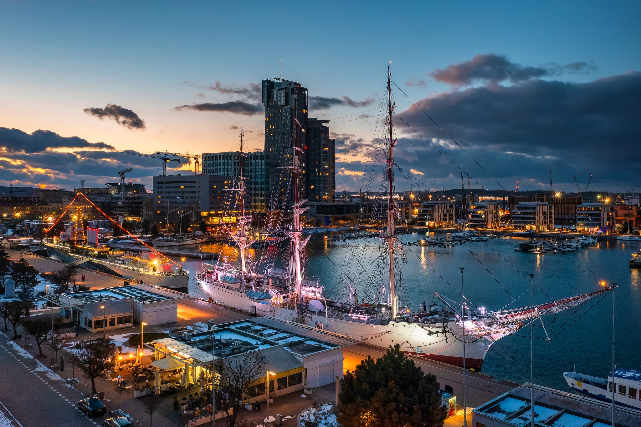 Nocny widok na port w Gdyni z żaglowcem Dar Pomorza na pierwszym planie i Sea Towers w tle, oświetlony ciepłym światłem.