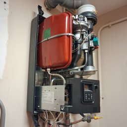 LK Serwis - Zamontowany na ścianie piec gazowy z czerwonym zbiornikiem i szarym kominem. Widoczne rury, kable i panel sterowania. Ujęcie z bliska, oświetlenie naturalne.
