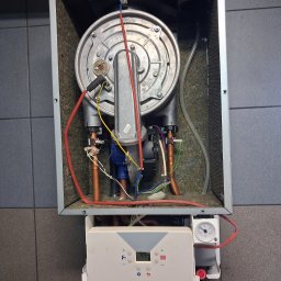 LK Serwis - Otwarty piec gazowy z widocznymi elementami wewnętrznymi, w tym palnikiem, przewodami i elektroniką. Widoczny manometr i panel sterowania. Tło stanowią szare płytki.