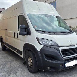 Transport Marcin Augustyniak - Biały bus dostawczy Peugeot zaparkowany na brukowanej posesji, widok z przodu, szara kamienna barierka w tle, pochmurne niebo.