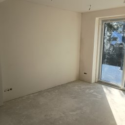 Janusz Woźniak - Pokój po szpachlowaniu i malowaniu na biało, widoczne gniazdka elektryczne, surowa podłoga i okno z widokiem na zewnątrz. W tle wystające kable z sufitu.