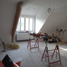 Janusz Woźniak - Poddasze w trakcie prac wykończeniowych: widoczne narzędzia na kozłach, przewody, okno dachowe i drewniana belka konstrukcyjna. Ściany przygotowane do malowania.
