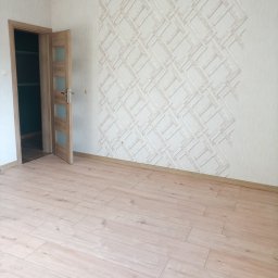 Janusz Woźniak - Jasne panele podłogowe w pokoju z otwartymi, drewnianymi drzwiami i ścianą z wzorzystą tapetą w geometryczne romby. Widok na wnętrze po remoncie.