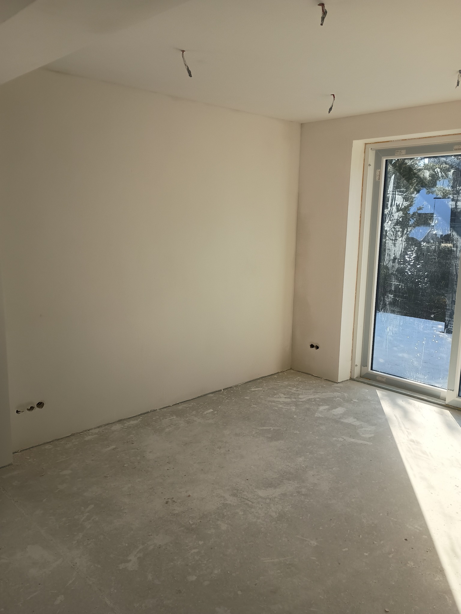 Pokój po szpachlowaniu i malowaniu na biało, widoczne gniazdka elektryczne, surowa podłoga i okno z widokiem na zewnątrz. W tle wystające kable z sufitu.
