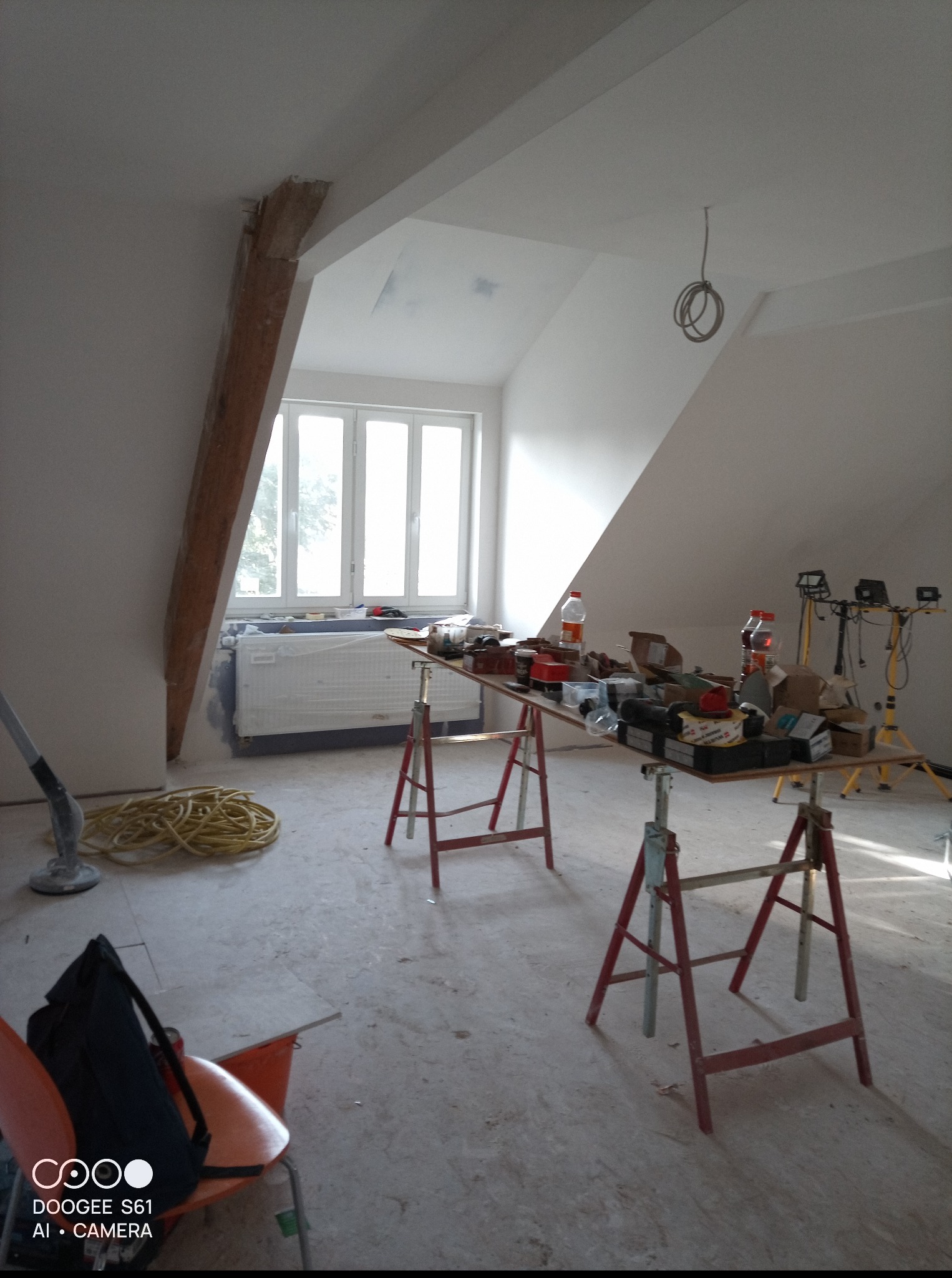 Poddasze w trakcie prac wykończeniowych: widoczne narzędzia na kozłach, przewody, okno dachowe i drewniana belka konstrukcyjna. Ściany przygotowane do malowania.