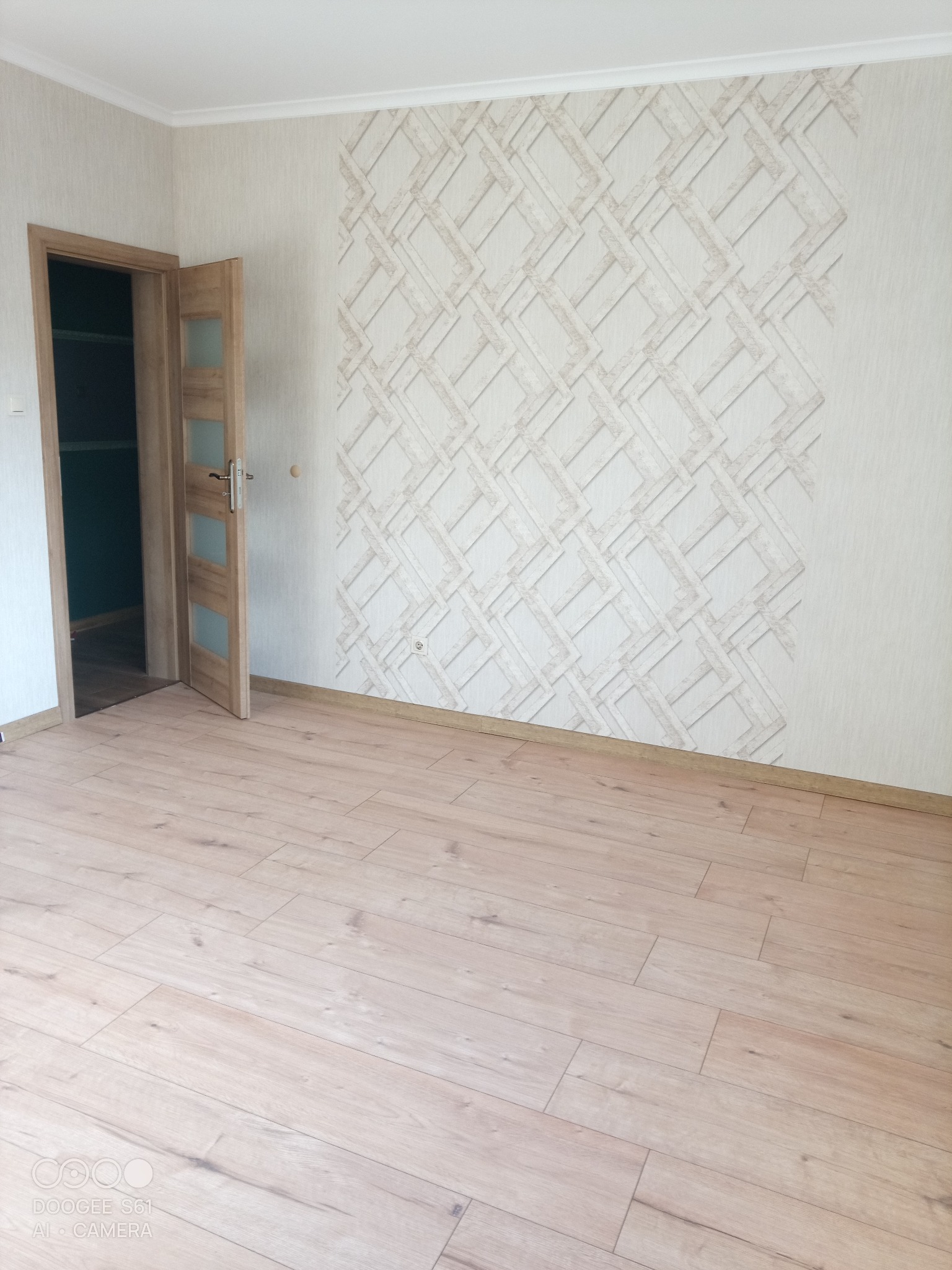 Jasne panele podłogowe w pokoju z otwartymi, drewnianymi drzwiami i ścianą z wzorzystą tapetą w geometryczne romby. Widok na wnętrze po remoncie.
