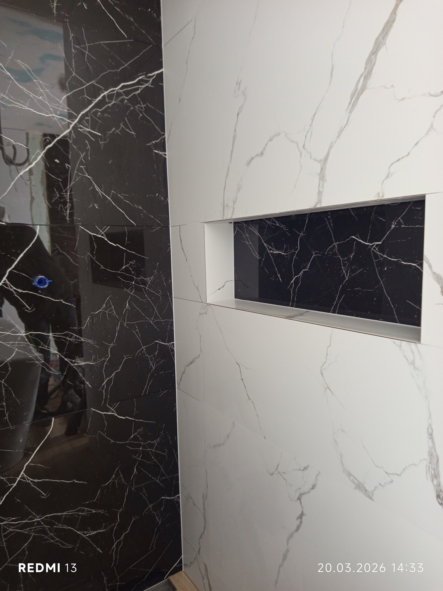 Wykończona ściana łazienki z czarno-białych płytek imitujących marmur. W białej części wnęka wyłożona czarną płytką. Widoczne fugi i precyzyjne ułożenie płytek.