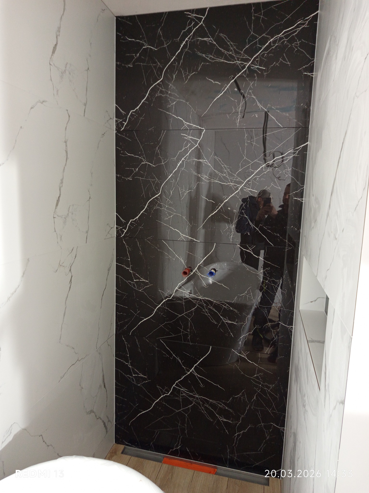 Wykończona ściana w łazience z czarnych i białych płytek imitujących marmur. Widoczne elementy instalacji wodnej i odbicie osoby robiącej zdjęcie.