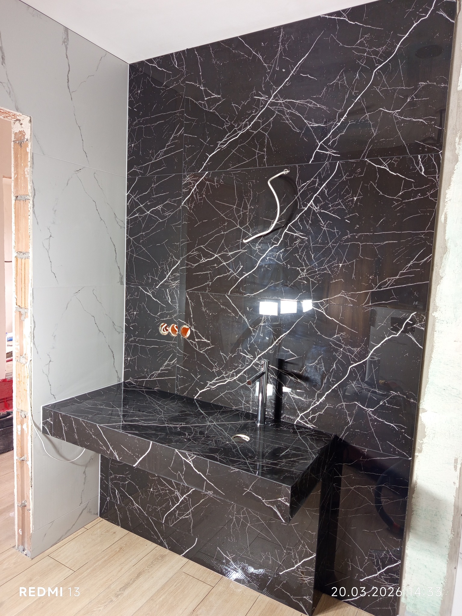 Częściowo wykończona łazienka z czarną marmurową ścianą i blatem pod umywalkę. Widoczne przyłącza wodne i fragment podłogi. Ujęcie z bliska, podkreślające detale.