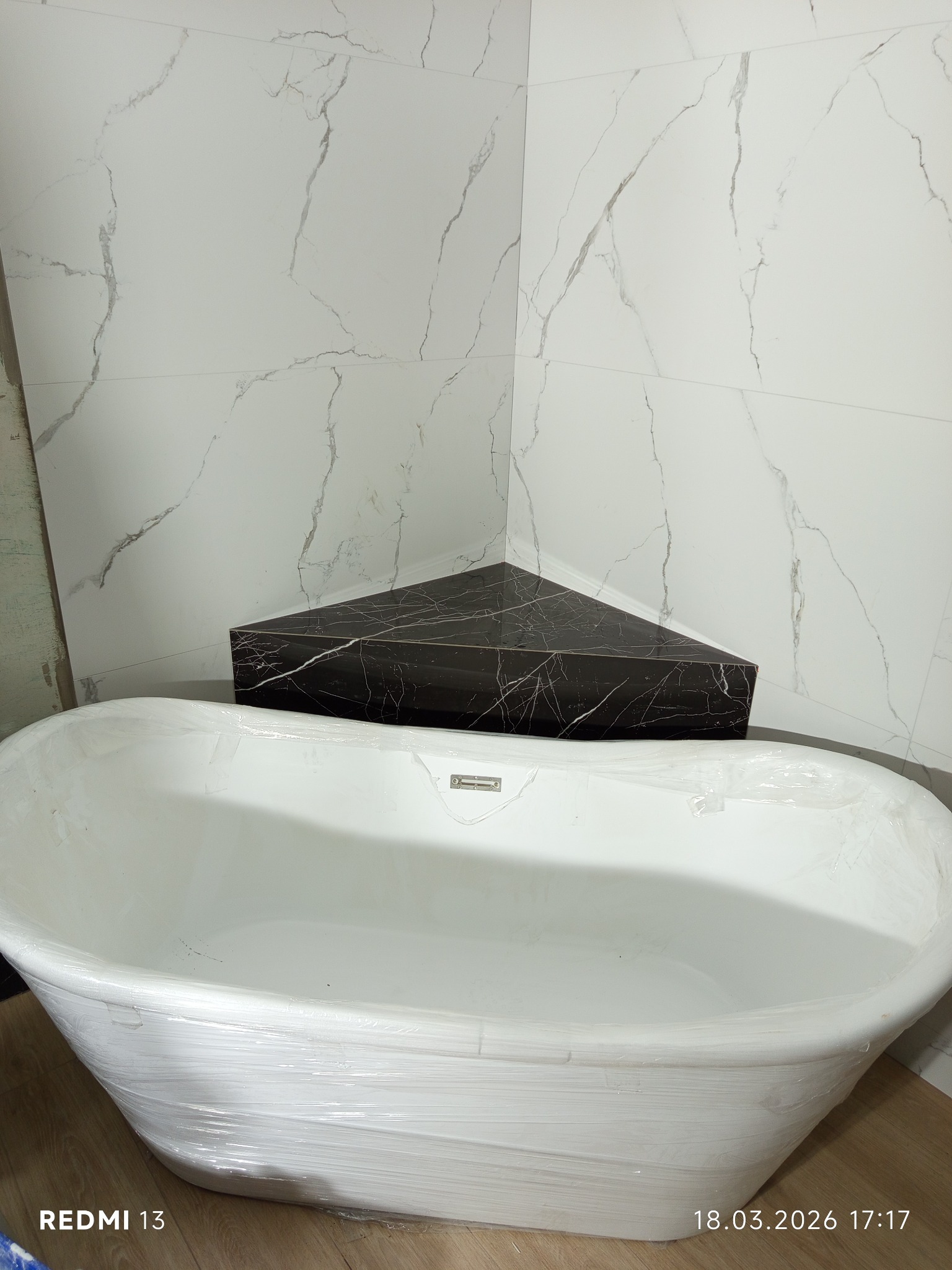 Biała wanna owinięta folią ochronną na tle ściany z białych płytek imitujących marmur i czarnej, marmurkowej półki narożnej. Widoczny fragment podłogi.