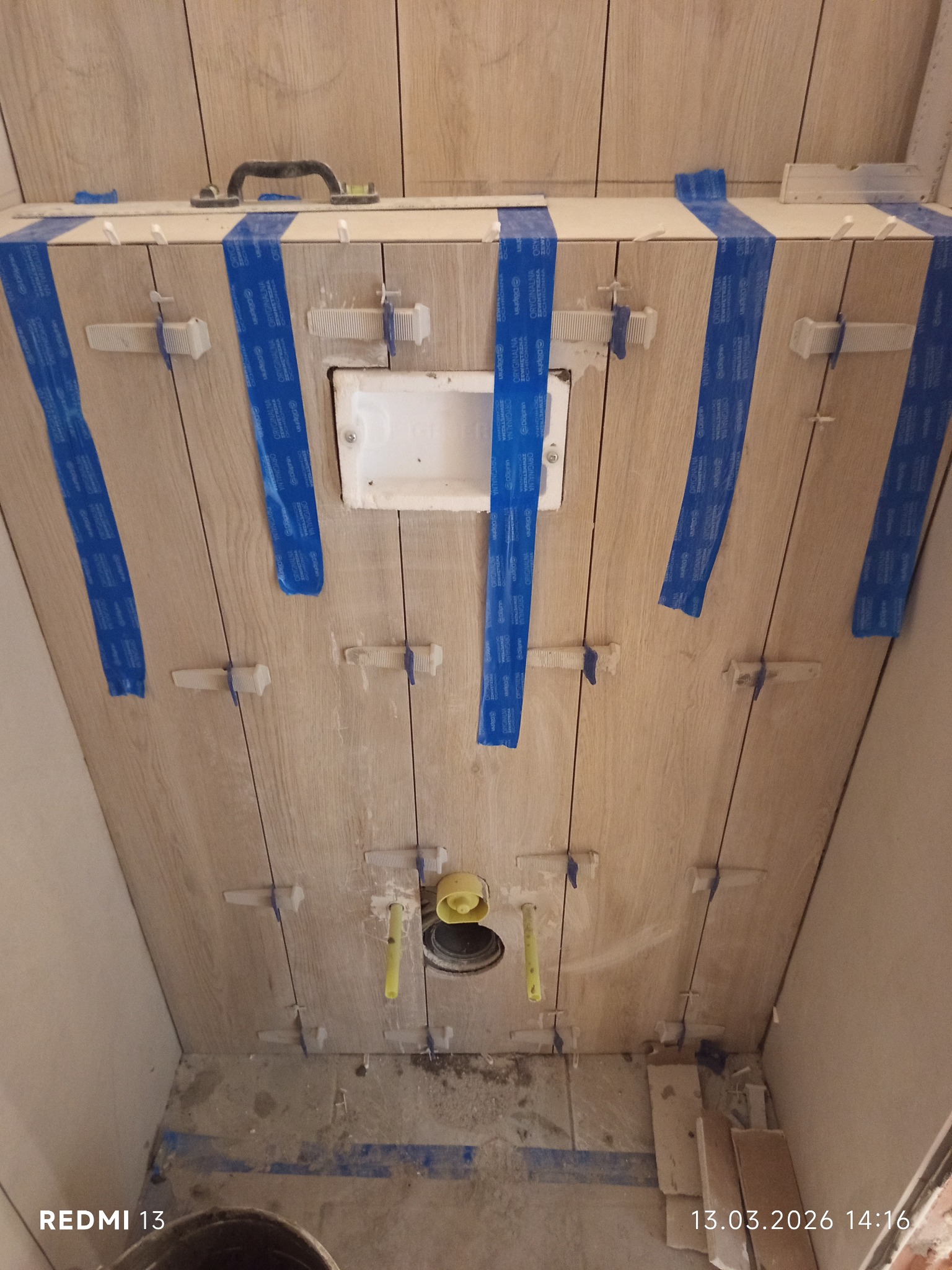 Montaż płytek imitujących drewno na ścianie w łazience. Widoczne krzyżyki dystansowe, taśma malarska i otwór na instalację. Prace glazurnicze w toku.