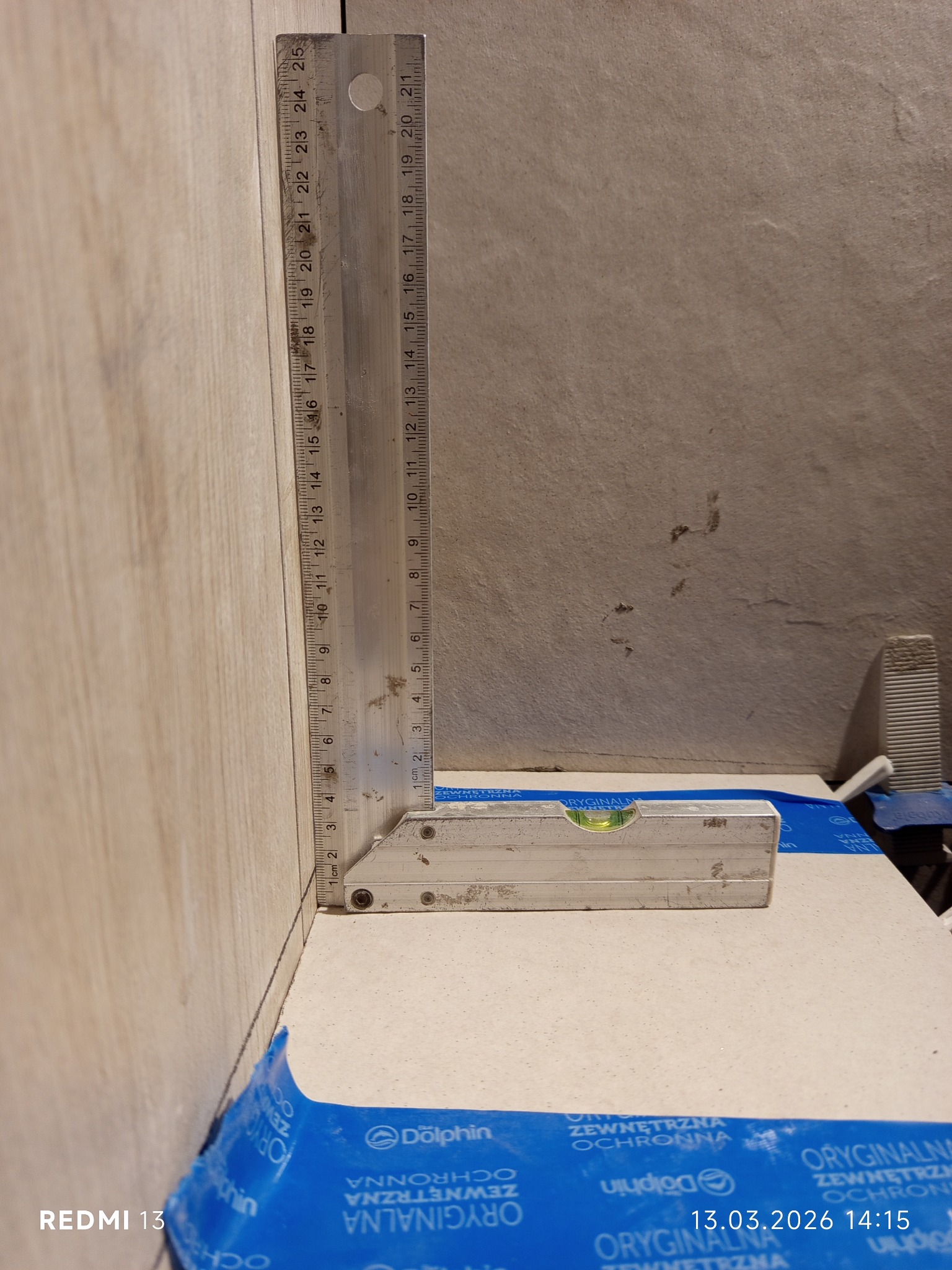 Pomiar kąta prostego przy użyciu kątownika na jasnej płytce, zabezpieczonej folią ochronną. W tle widoczna ściana i elementy montażowe.