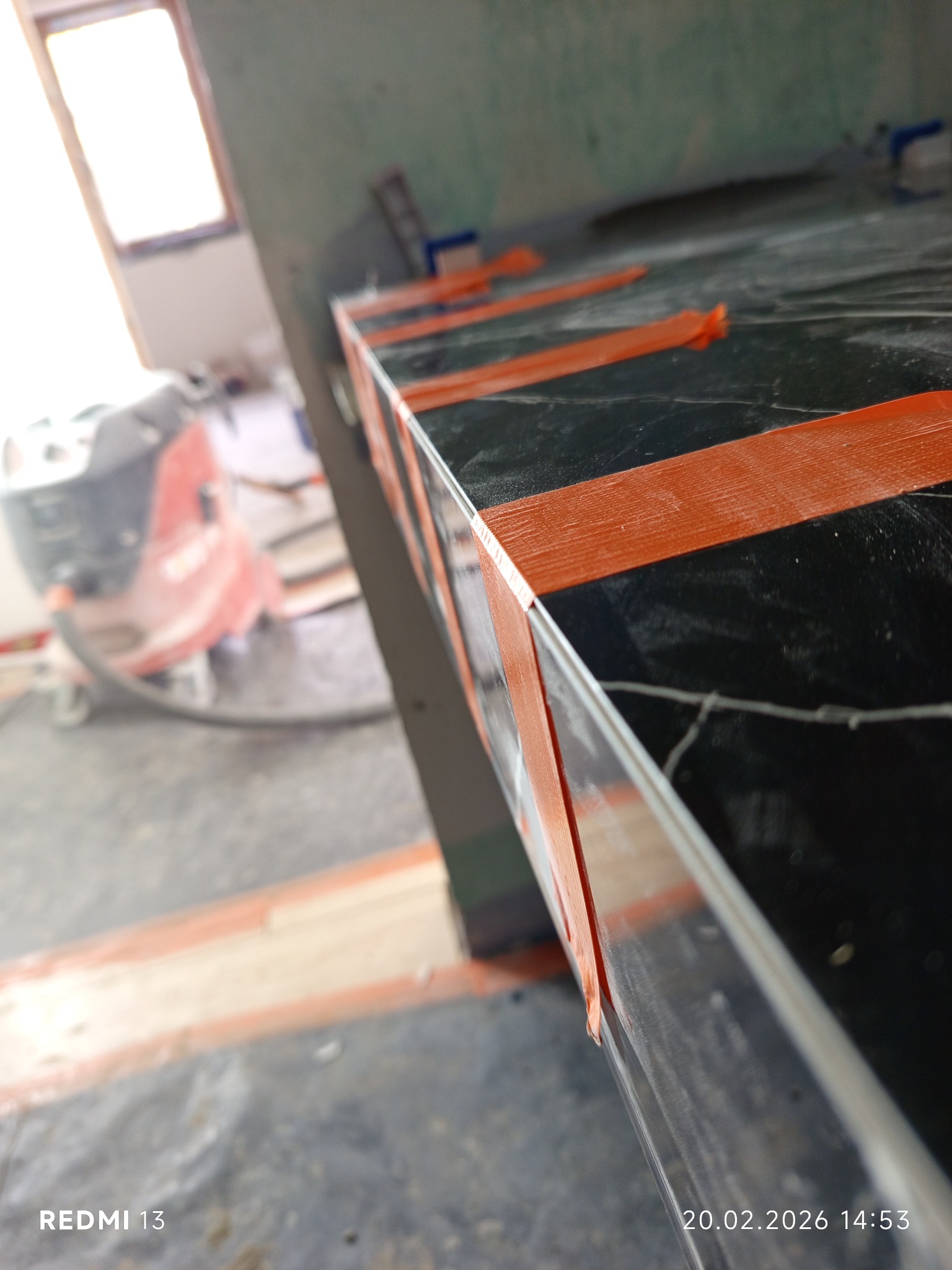 Czarny blat z marmurowym wzorem, zabezpieczony pomarańczową taśmą malarską, w trakcie prac wykończeniowych. W tle odkurzacz przemysłowy i okno.