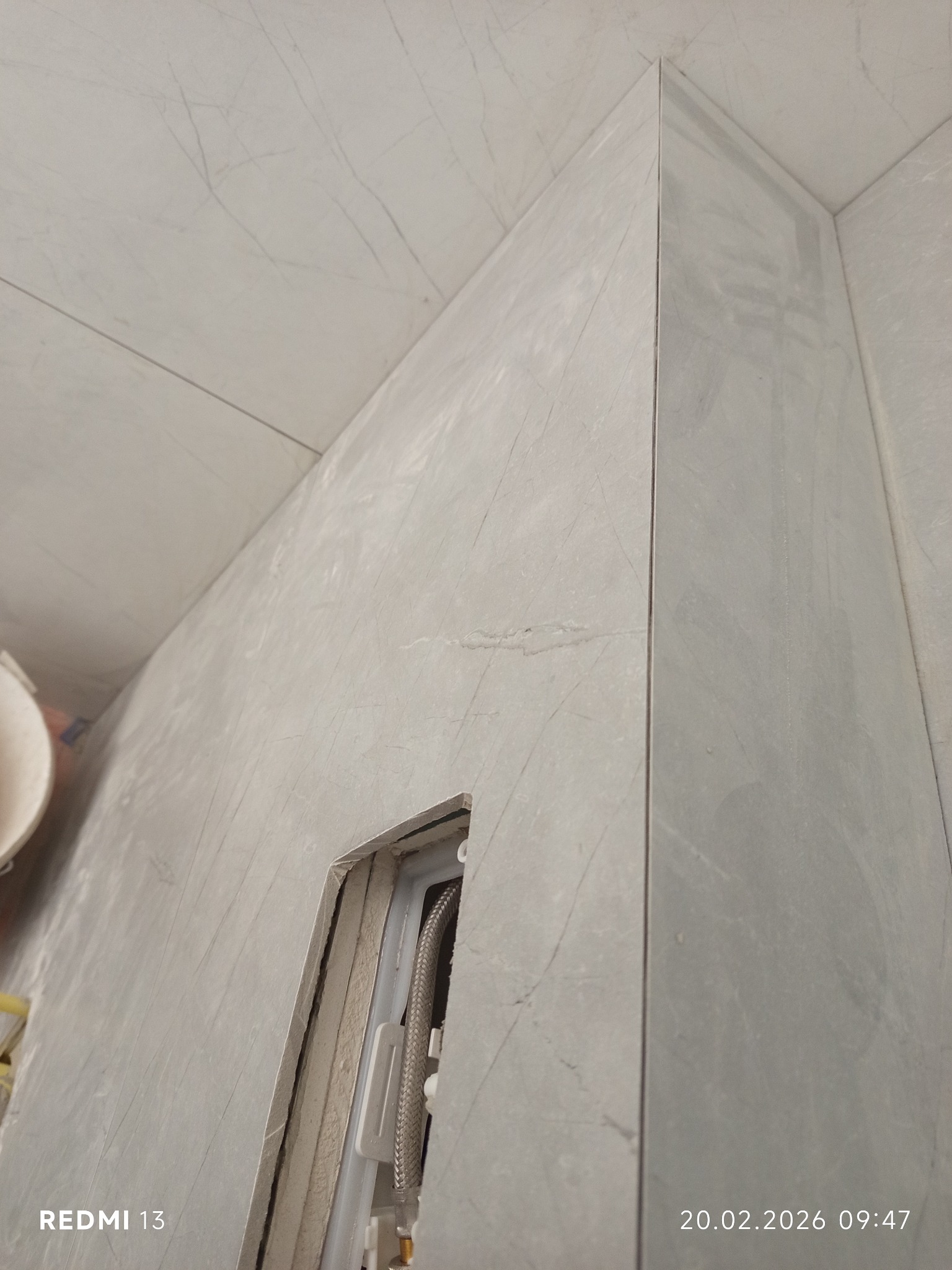 Wykończona ściana w łazience z szarymi płytkami imitującymi marmur. Widoczny fragment instalacji wodnej w otworze. Minimalistyczny design, precyzyjne ułożenie płytek.