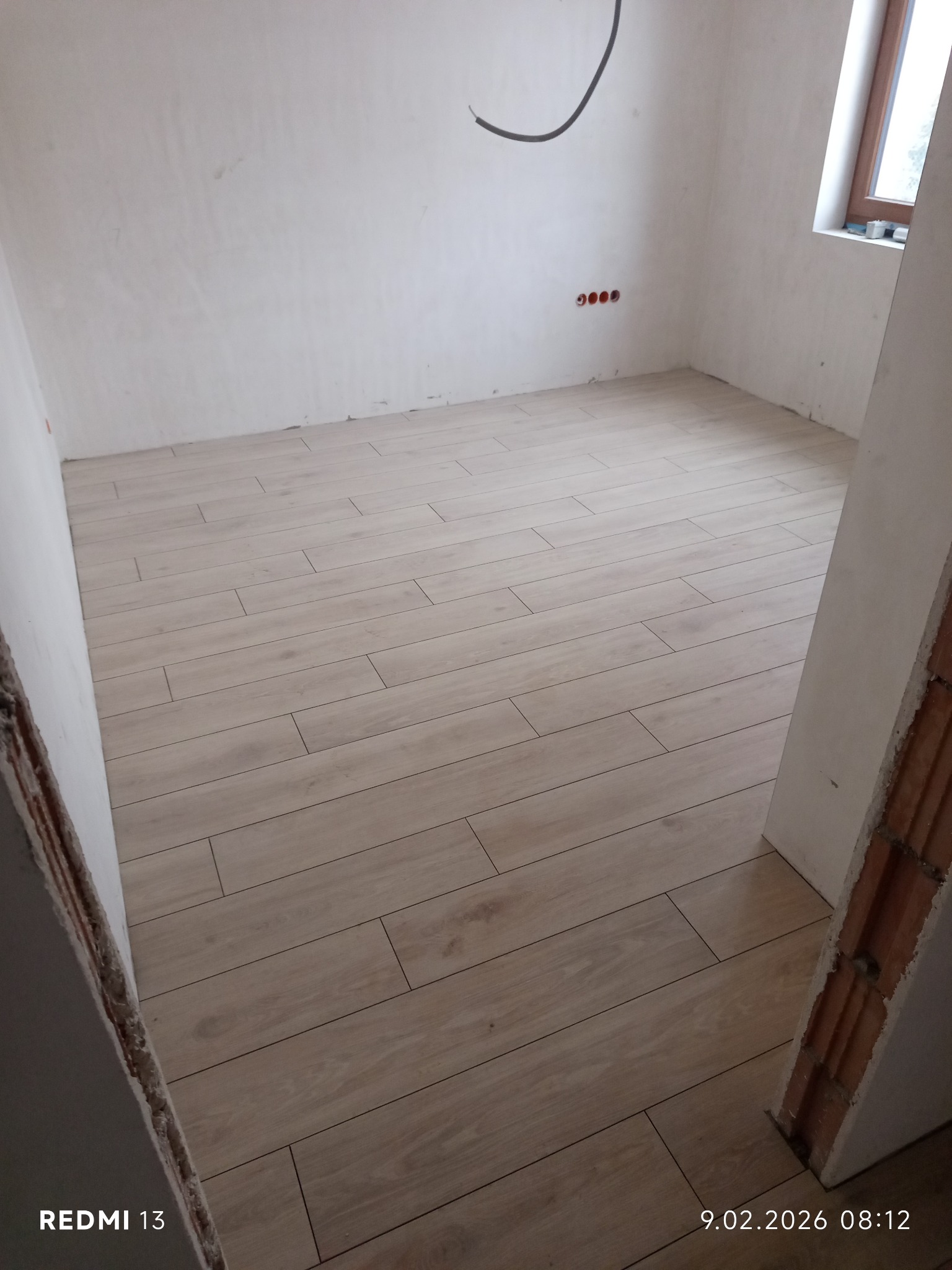 Ułożone panele podłogowe w jasnym odcieniu drewna w surowym wnętrzu. Widoczne gniazdka elektryczne i fragment okna. Świeżo wykończona podłoga w trakcie remontu.