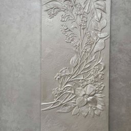 ARTESSA HOME SPÓŁKA Z OGRANICZONĄ ODPOWIEDZIALNOŚCIĄ - Dekoracyjny panel ścienny z gipsową płaskorzeźbą kwiatową w odcieniach szarości, umieszczony na ścianie o fakturze betonu. Subtelne światło podkreśla detale.