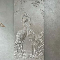 ARTESSA HOME SPÓŁKA Z OGRANICZONĄ ODPOWIEDZIALNOŚCIĄ - Dekoracyjna płaskorzeźba ścienna z motywem dwóch żurawi i roślinności, wykonana w jasnych odcieniach szarości, umieszczona na ścianie o fakturze betonu.