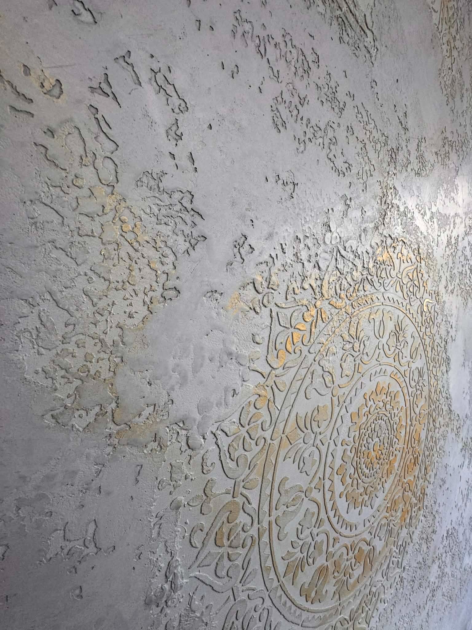 Dekoracyjna ściana z szarego tynku strukturalnego z ornamentem w stylu mandali, wykończonym złotą farbą. Efektowna faktura i unikalny wzór.