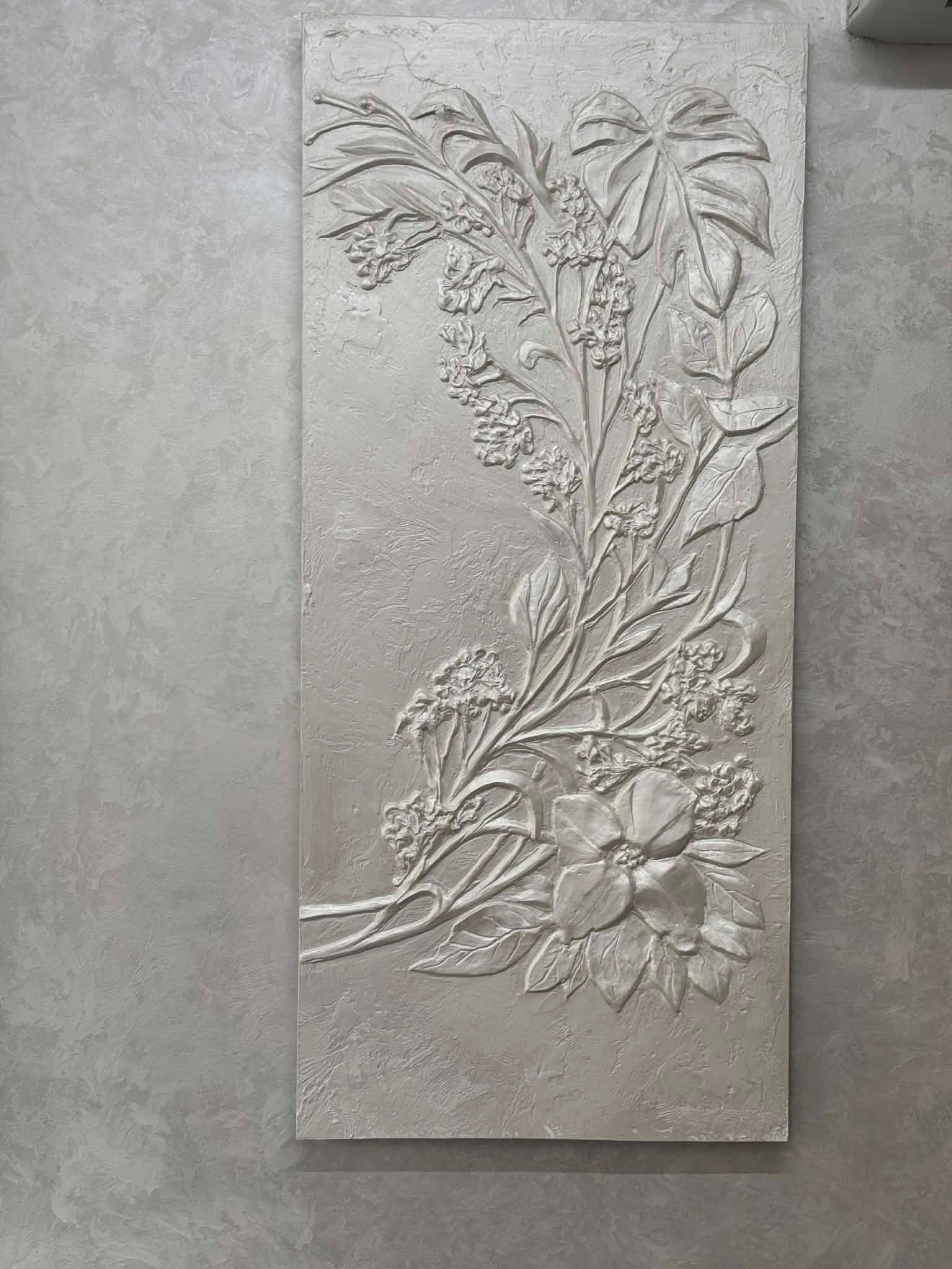 Dekoracyjny panel ścienny z gipsową płaskorzeźbą kwiatową w odcieniach szarości, umieszczony na ścianie o fakturze betonu. Subtelne światło podkreśla detale.