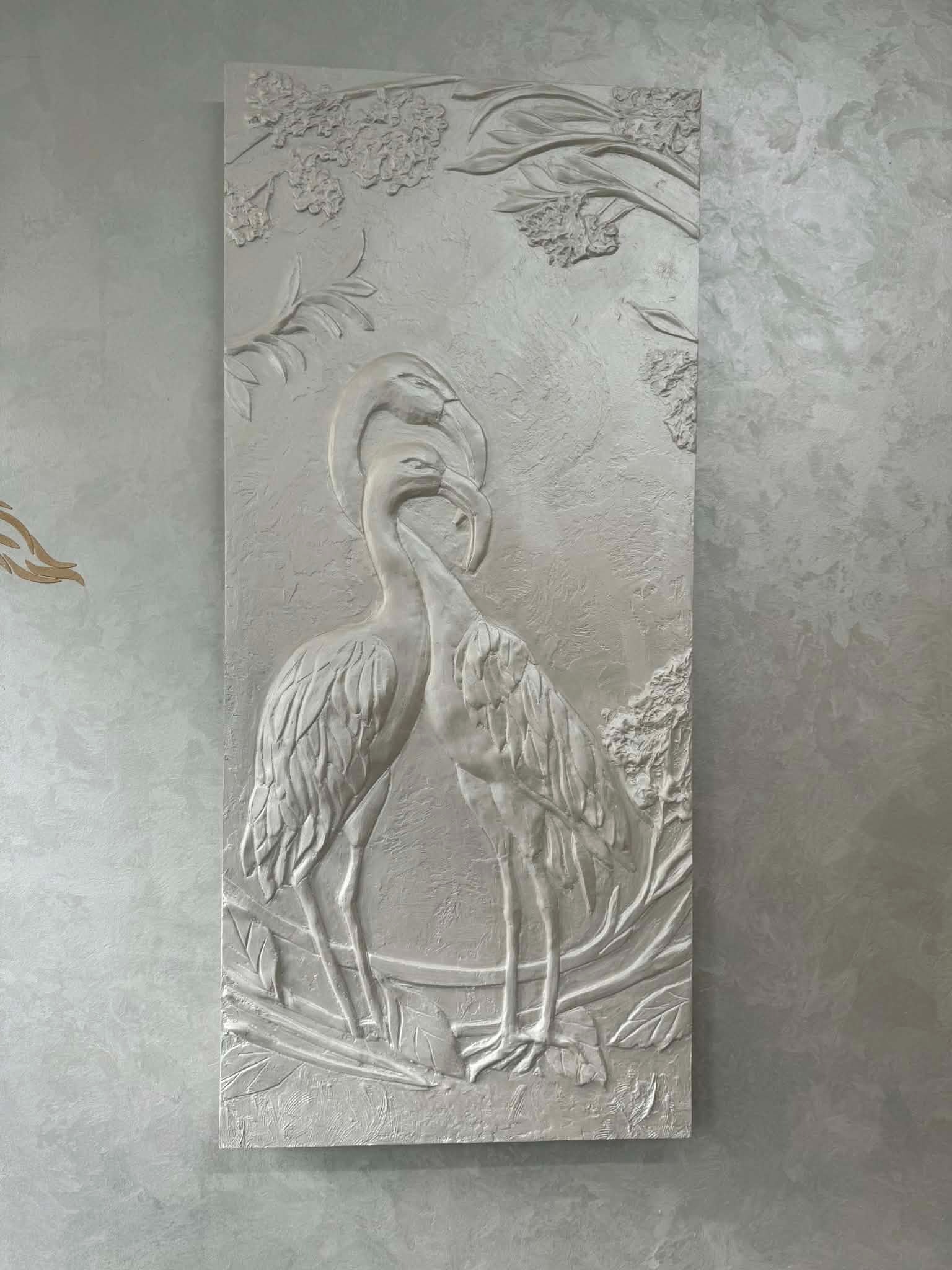 Dekoracyjna płaskorzeźba ścienna z motywem dwóch żurawi i roślinności, wykonana w jasnych odcieniach szarości, umieszczona na ścianie o fakturze betonu.