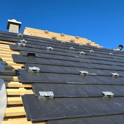 MKDienstleistungen Sp. z o.o. - Montaż paneli fotowoltaicznych na dachu z czarnej dachówki, widoczne metalowe uchwyty i fragment konstrukcji drewnianej. Błękitne niebo w tle.