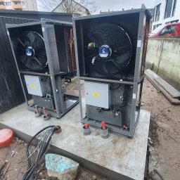 MKDienstleistungen Sp. z o.o. - Dwie szare pompy ciepła powietrze-woda na betonowej podstawie, z widocznymi wentylatorami i elementami instalacji. W tle budynek mieszkalny i czerwony samochód dostawczy.