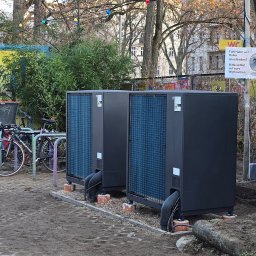 MKDienstleistungen Sp. z o.o. - Dwie czarne pompy ciepła na zewnątrz, z widocznymi rurami i elementami instalacji, na tle placu zabaw z rowerami i zielenią. Nietypowa lokalizacja.