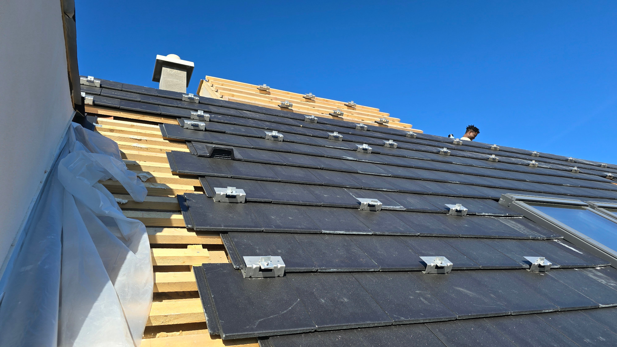 Montaż paneli fotowoltaicznych na dachu z czarnej dachówki, widoczne metalowe uchwyty i fragment konstrukcji drewnianej. Błękitne niebo w tle.
