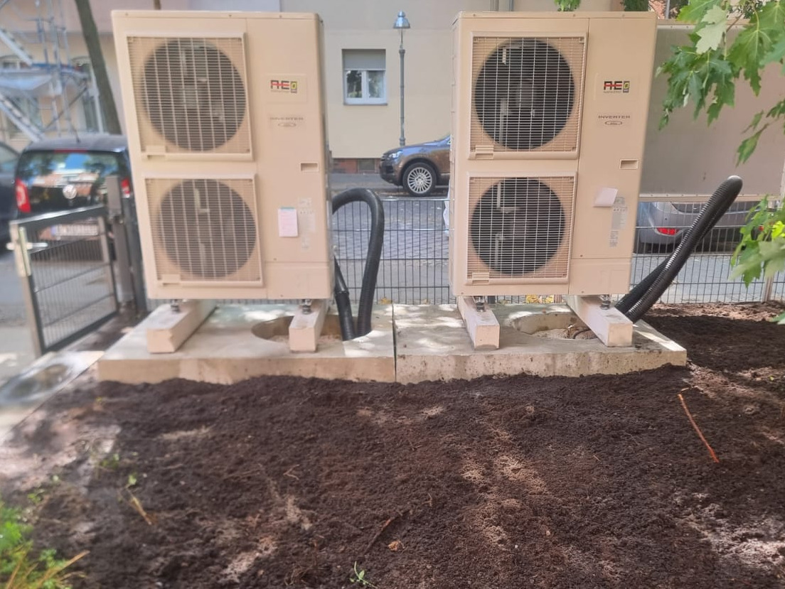 Dwie jednostki zewnętrzne klimatyzacji typu split, ustawione na betonowych podstawach w ogrodzie, z widocznymi przewodami i świeżo przekopaną ziemią wokół.