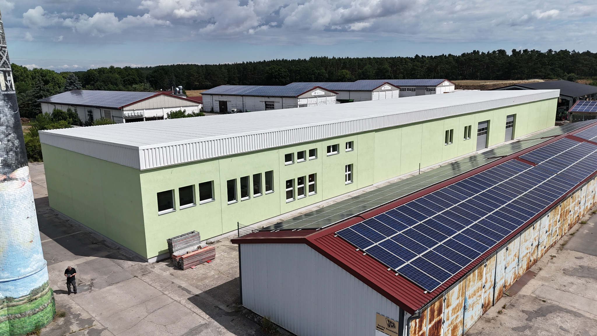 Zielony budynek z panelami słonecznymi na dachu, widok z góry. W tle inne budynki z panelami i las. Na pierwszym planie komin z malowidłami.