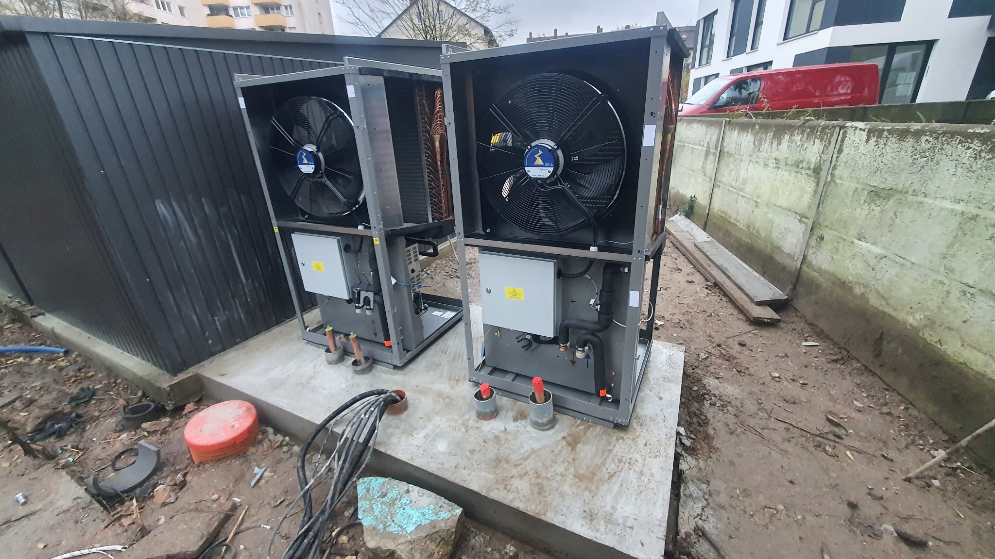 Dwie szare pompy ciepła powietrze-woda na betonowej podstawie, z widocznymi wentylatorami i elementami instalacji. W tle budynek mieszkalny i czerwony samochód dostawczy.