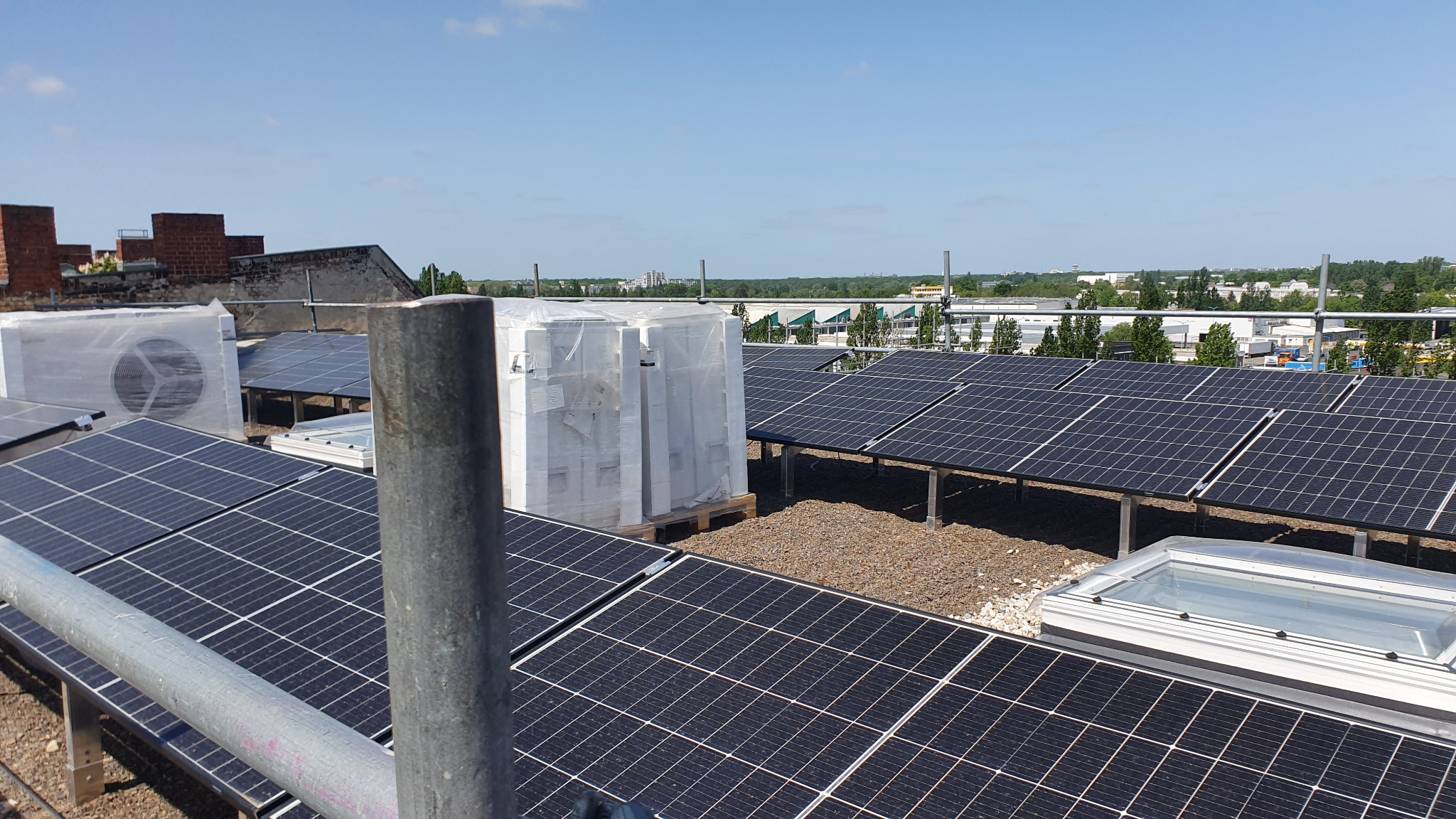Instalacja fotowoltaiczna na dachu budynku, widoczne panele i elementy montażowe. W tle zabudowa miejska i błękitne niebo. Część urządzeń jeszcze w folii ochronnej.