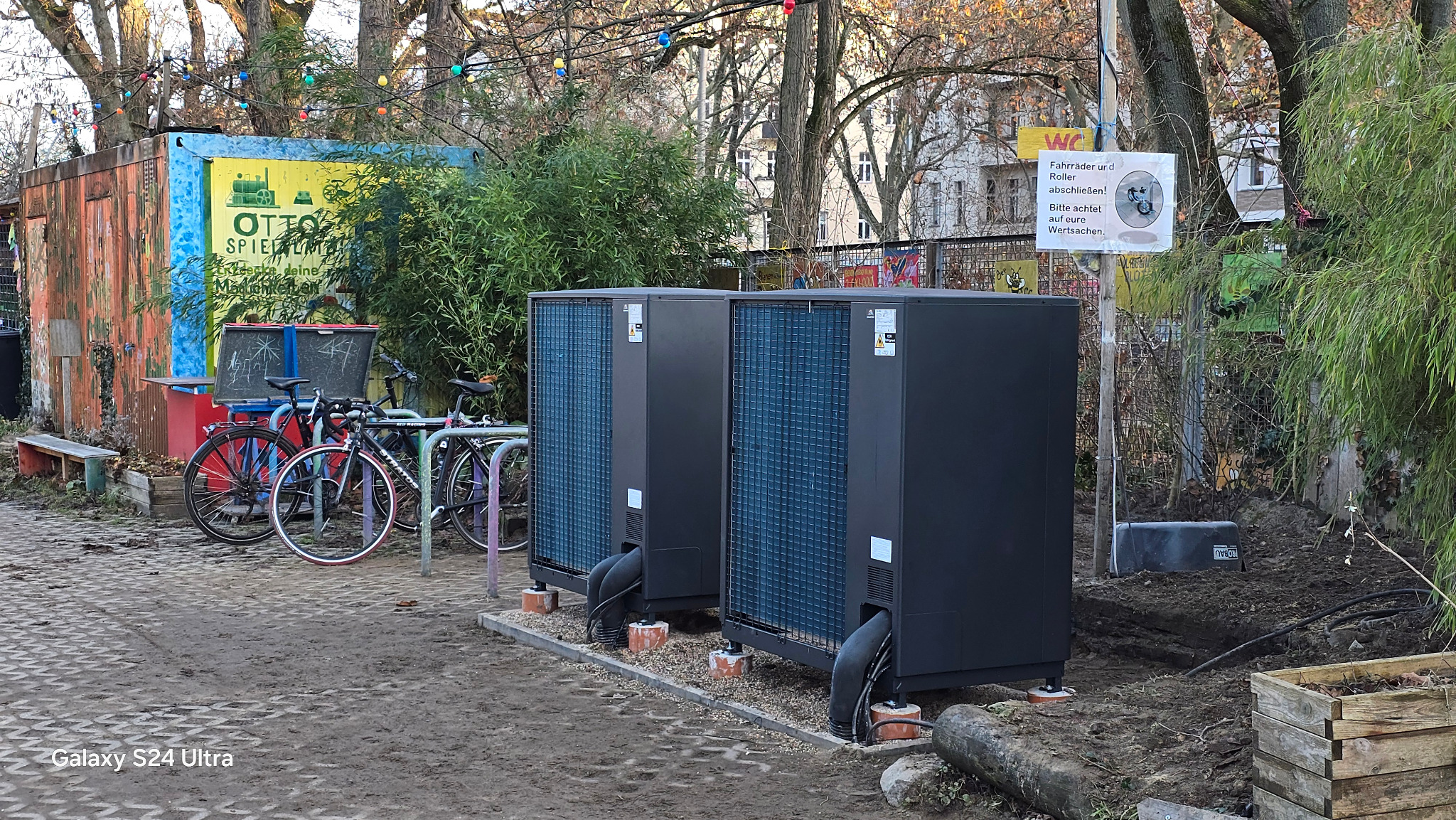 Dwie czarne pompy ciepła na zewnątrz, z widocznymi rurami i elementami instalacji, na tle placu zabaw z rowerami i zielenią. Nietypowa lokalizacja.