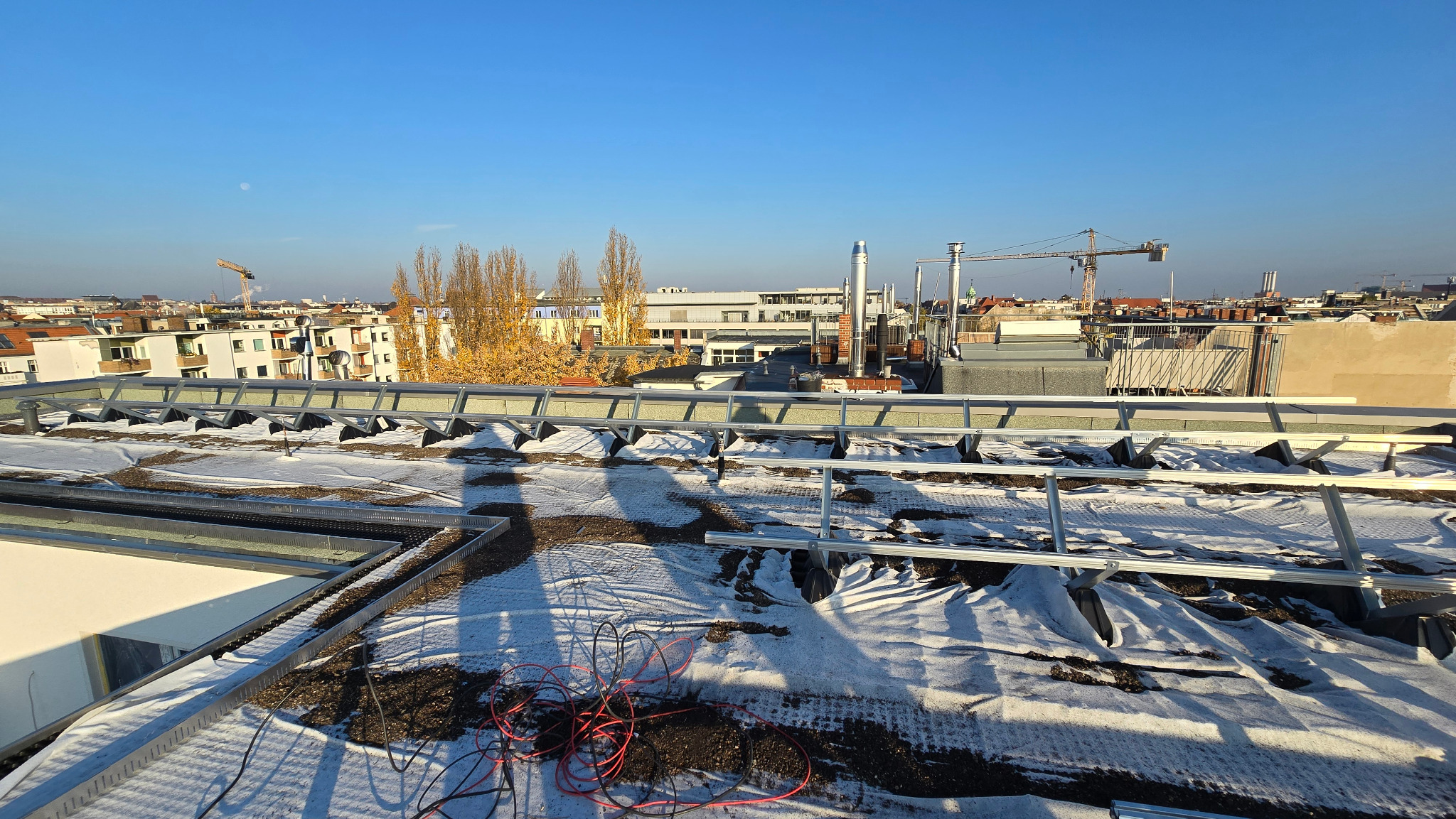 Montaż konstrukcji wsporczej pod panele fotowoltaiczne na dachu budynku w słoneczny dzień. Widoczne kable i elementy montażowe. W tle zabudowa miejska i żurawie budowlane.
