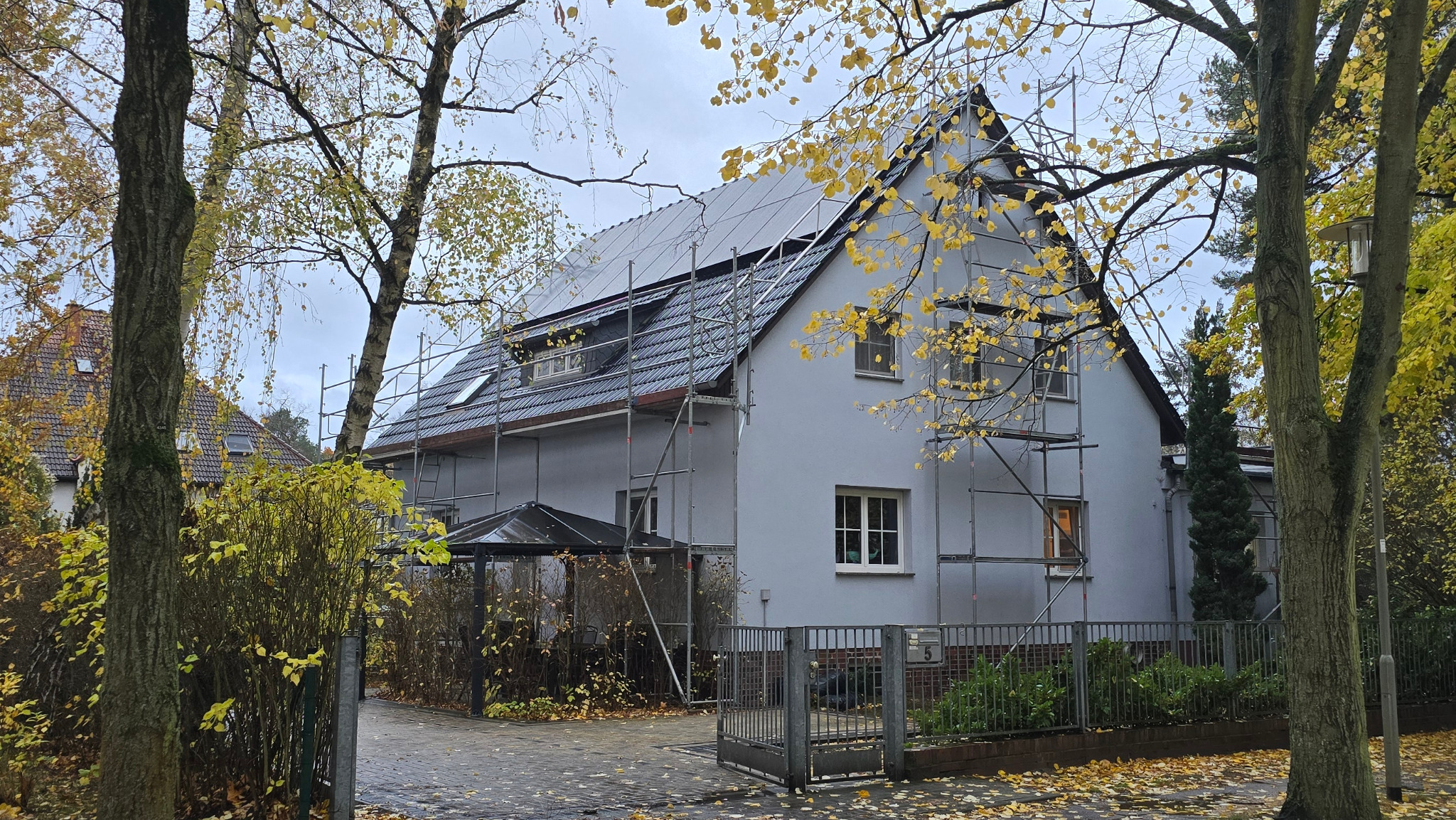 Dom jednorodzinny z panelami słonecznymi na dachu, otoczony rusztowaniem. Jesienne liście na drzewach i chodniku. Widoczny fragment ogrodzenia i wejścia.